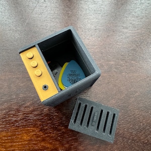 May include: A small, black and yellow 3D printed box with a removable lid. The box is open and contains a few guitar picks and a small black object. The box has a series of small, round, yellow buttons on the front.