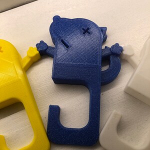 May include: Three 3D printed door stops in the shape of a cartoon character. The door stops are blue, yellow, and white. Each door stop has a hook on the bottom to hold a door open.