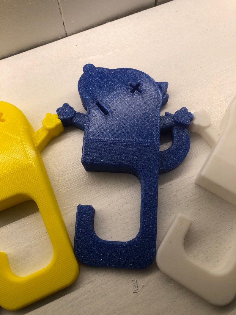 May include: Three 3D printed door stops in the shape of a cartoon character. The door stops are blue, yellow, and white. Each door stop has a hook on the bottom to hold a door open.