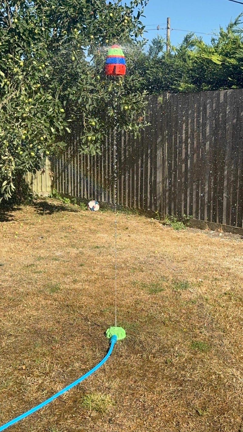 May include: A colorful sprinkler sprays water in a backyard. The sprinkler is red, blue, green, and yellow. A soccer ball sits on the dry grass. A blue hose connects to the sprinkler base.