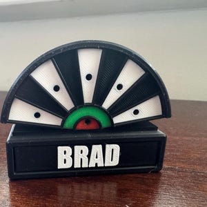 May include: Black and white target with a green and red bullseye. The target is mounted on a black base with the name "BRAD" printed in white.
