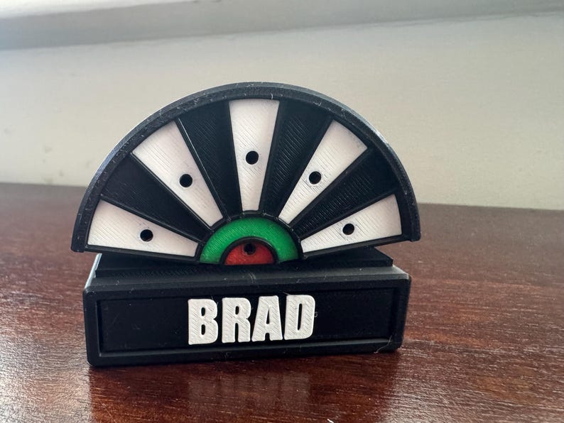 May include: Black and white target with a green and red bullseye. The target is mounted on a black base with the name "BRAD" printed in white.