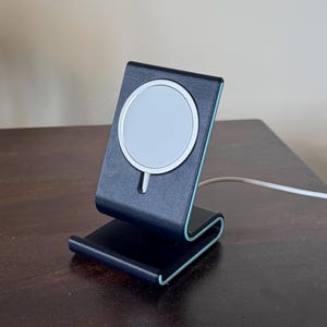 May include: A black and teal wireless charging stand with a white charging surface. The stand is made of a curved design with a teal accent. A white charging cable is connected to the stand.
