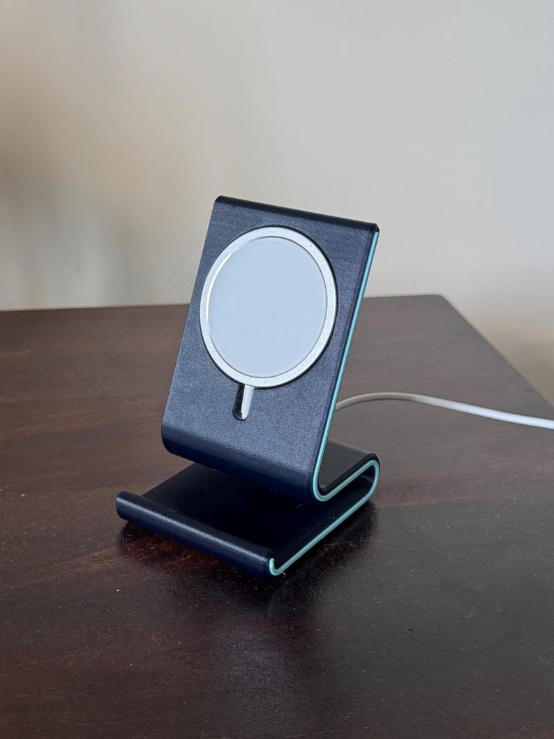 May include: A black and teal wireless charging stand with a white charging surface. The stand is made of a curved design with a teal accent. A white charging cable is connected to the stand.