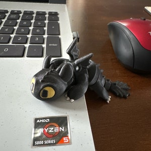 May include: A black 3D printed figurine of a dragon with yellow eyes. The dragon is sitting on a laptop with a sticker that says "AMD Ryzen 5 5000 Series".