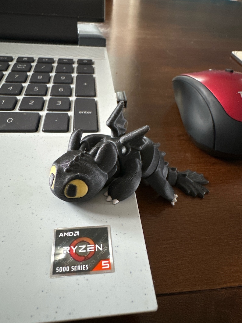 May include: A black 3D printed figurine of a dragon with yellow eyes. The dragon is sitting on a laptop with a sticker that says "AMD Ryzen 5 5000 Series".