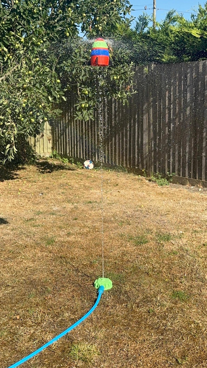 May include: A backyard scene featuring a colorful water sprinkler spraying water. A soccer ball sits on the dry grass. A wooden fence and a tree with green apples are in the background. The sprinkler is connected to a blue hose.