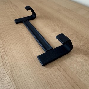May include: A black, 3D-printed, two-sided hanger with a rectangular base and curved hooks. The hanger is designed to hold items and is placed on a light-colored wooden surface.