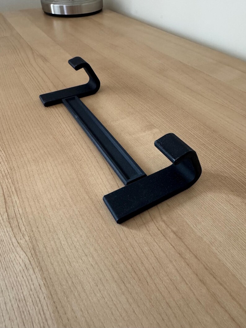 May include: A black, 3D-printed, two-sided hanger with a rectangular base and curved hooks. The hanger is designed to hold items and is placed on a light-colored wooden surface.
