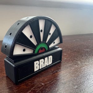 May include: A black and white dartboard with a green and red bullseye. The dartboard is mounted on a black base with the name "BRAD" printed in white letters.