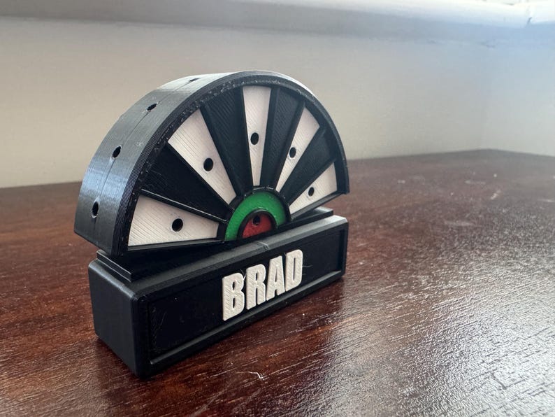 May include: A black and white dartboard with a green and red bullseye. The dartboard is mounted on a black base with the name "BRAD" printed in white letters.