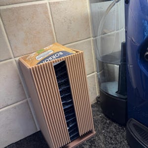 May include: A light brown, ribbed tea bag holder with a box of tea bags inside. The holder is rectangular and sits on a countertop next to a coffee maker. The tea bag box has the word "BORBON" printed on it.