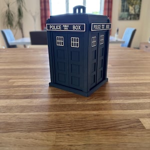 May include: A dark blue Police Box replica with white lettering and window details. The text "POLICE PUBLIC CALL BOX" is displayed on the top. The box is a miniature model, likely a collectible or decorative item.
