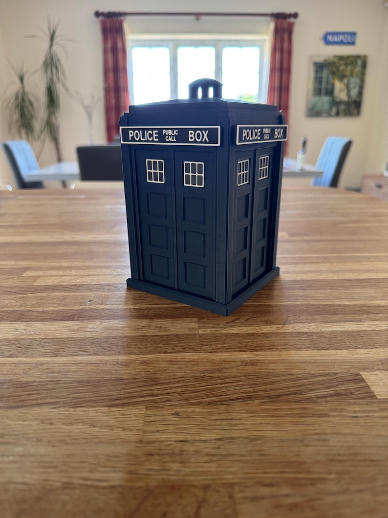 May include: A dark blue Police Box replica with white lettering and window details. The text "POLICE PUBLIC CALL BOX" is displayed on the top. The box is a miniature model, likely a collectible or decorative item.