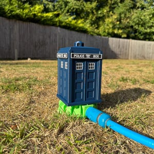 May include: A blue Police Box garden hose holder with white lettering and window details. The holder is connected to a blue hose and sits on a green base. The scene is set outdoors on a sunny day.