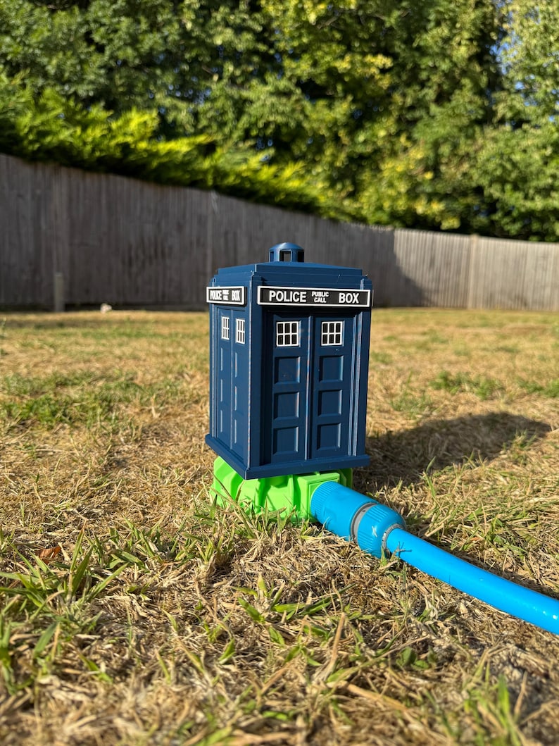 May include: A blue Police Box garden hose holder with white lettering and window details. The holder is connected to a blue hose and sits on a green base. The scene is set outdoors on a sunny day.