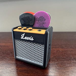 May include: A black and yellow miniature guitar amplifier with a silver speaker grille. The amplifier has the word "Lewis" printed on the front. There are three knobs on the top of the amplifier and a compartment on the top for guitar picks. The picks are purple and black.