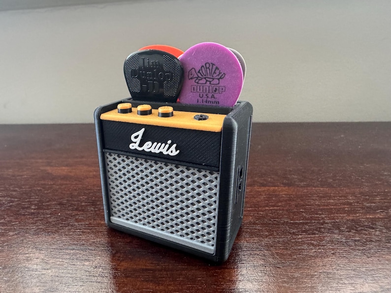 May include: A black and yellow miniature guitar amplifier with a silver speaker grille. The amplifier has the word "Lewis" printed on the front. There are three knobs on the top of the amplifier and a compartment on the top for guitar picks. The picks are purple and black.