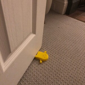 May include: A yellow doorstop shaped like a cartoon character with a simple face. The doorstop is lying on its side on a beige carpet.