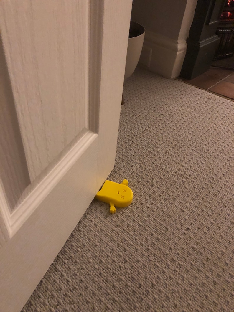 May include: A yellow doorstop shaped like a cartoon character with a simple face. The doorstop is lying on its side on a beige carpet.