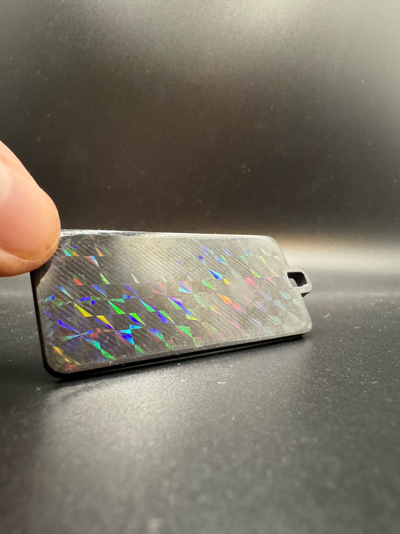 May include: A black rectangular tag with a holographic iridescent pattern. The tag has a small hole at the top for attaching a string or ribbon.