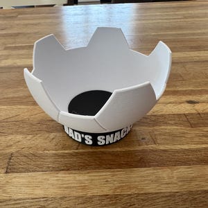 Op de afbeelding: Een witte, voetbalvormige kom met een zwarte basis. De basis heeft de tekst "DAD'S SNACKS" in het wit. De kom staat op een houten tafel.
