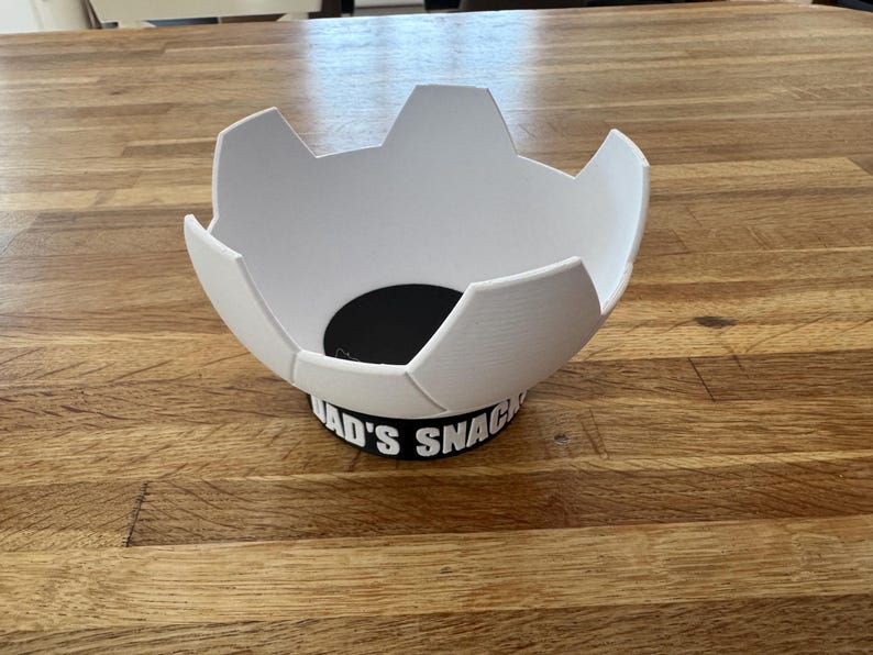 Op de afbeelding: Een witte, voetbalvormige kom met een zwarte basis. De basis heeft de tekst "DAD'S SNACKS" in het wit. De kom staat op een houten tafel.