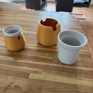 May include: Three decorative ceramic containers in shades of yellow and gray sit on a wooden surface. The containers have rounded shapes, with one featuring a unique cutout. The containers are of varying sizes, offering a modern aesthetic.