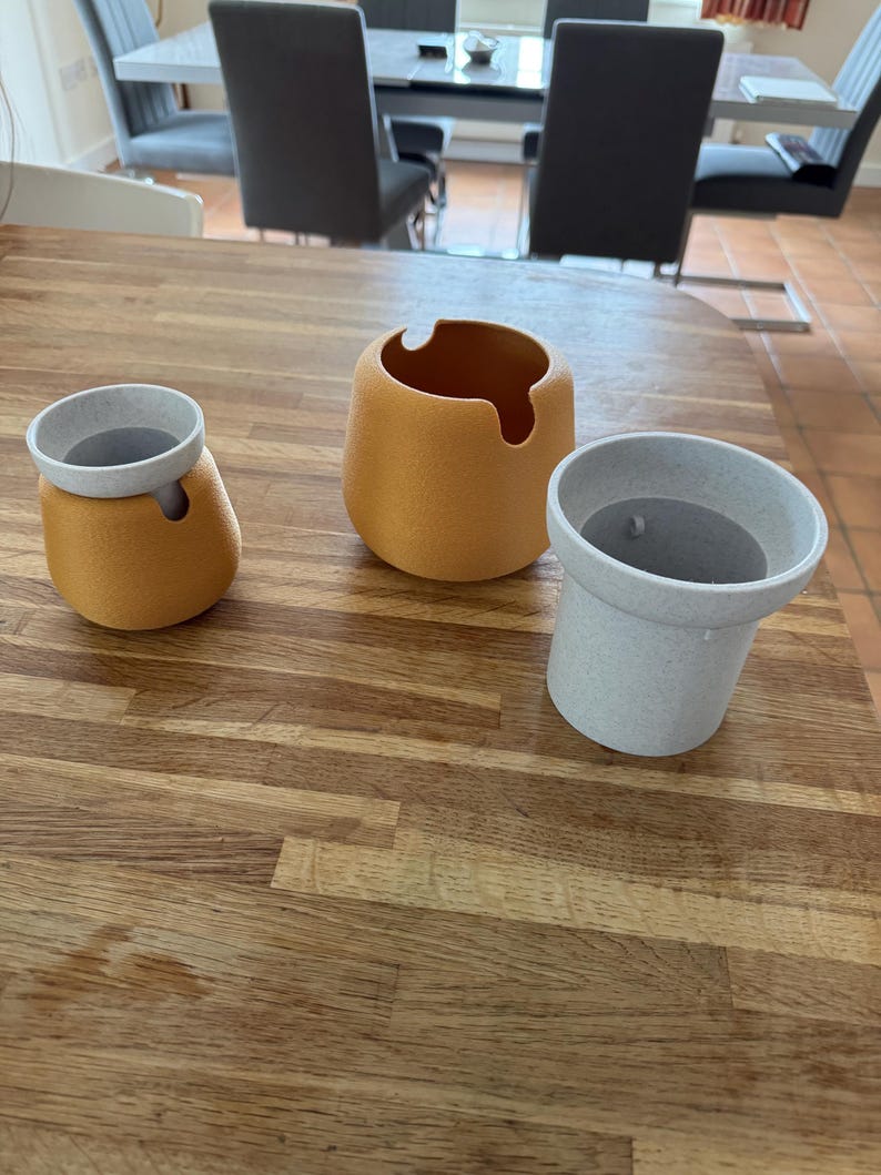 May include: Three decorative ceramic containers in shades of yellow and gray sit on a wooden surface. The containers have rounded shapes, with one featuring a unique cutout. The containers are of varying sizes, offering a modern aesthetic.