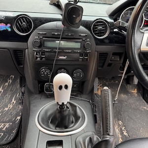 Op de afbeelding: Auto-interieur met een zwart dashboard en een witte, spookvormige versnellingspookknop. Het dashboard heeft een radio, ventilatieopeningen en een telefoonhouder. Een Coca-Cola-blikje hangt aan de achteruitkijkspiegel.