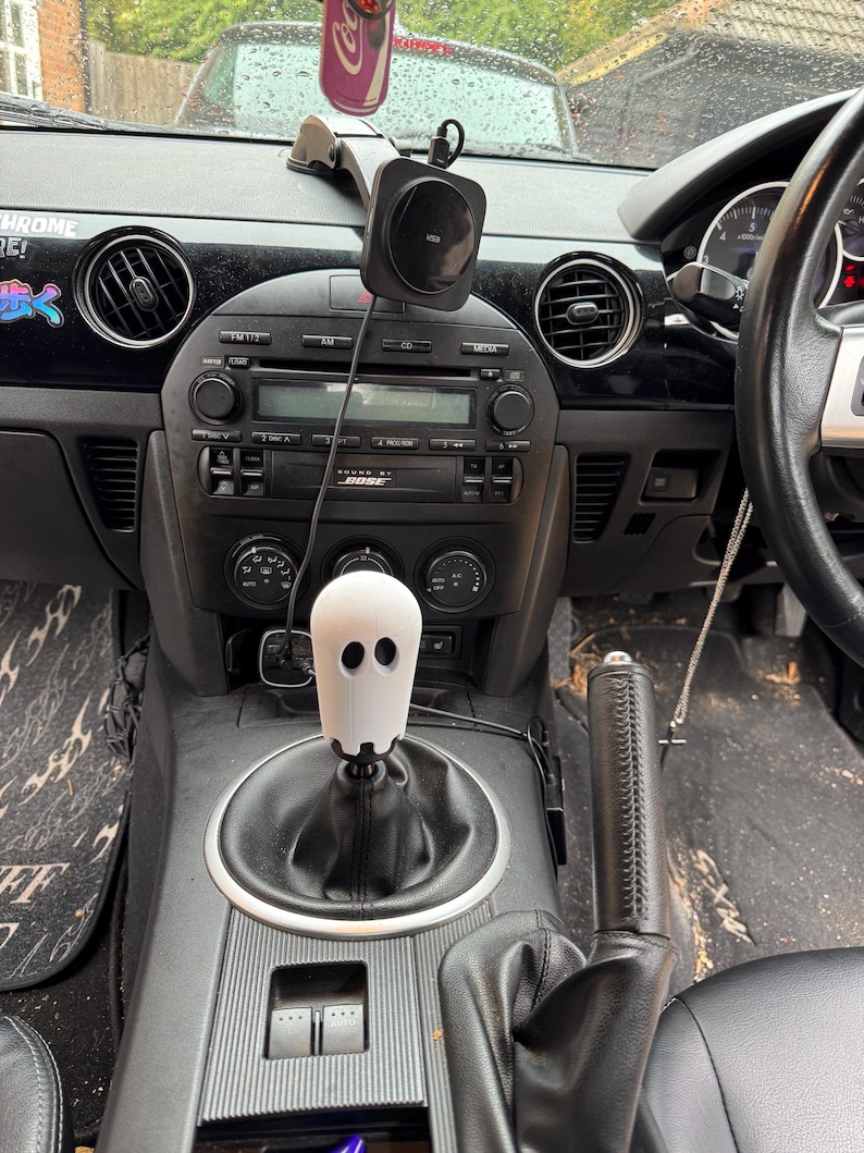 Op de afbeelding: Auto-interieur met een zwart dashboard en een witte, spookvormige versnellingspookknop. Het dashboard heeft een radio, ventilatieopeningen en een telefoonhouder. Een Coca-Cola-blikje hangt aan de achteruitkijkspiegel.