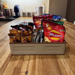 May include: A beige rectangular organizer labeled "CHOC BARS" and "SNACKS" filled with individually wrapped chocolate bars and bags of chips. The chips are in red, green, and blue packaging. The organizer sits on a wooden countertop.