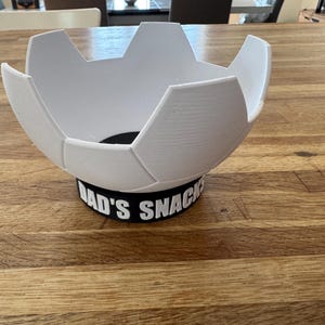 Op de afbeelding: Een witte, voetbalvormige kom met een zwarte basis. De basis heeft de tekst "DAD'S SNACKS" in het wit. De kom staat op een houten tafel.