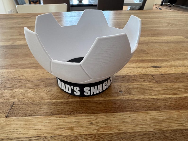 Op de afbeelding: Een witte, voetbalvormige kom met een zwarte basis. De basis heeft de tekst "DAD'S SNACKS" in het wit. De kom staat op een houten tafel.