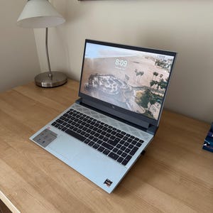 May include: A light blue laptop with a black keyboard is open on a wooden desk. The screen displays a beach scene with palm trees. A silver desk lamp and a decorative map are in the background.
