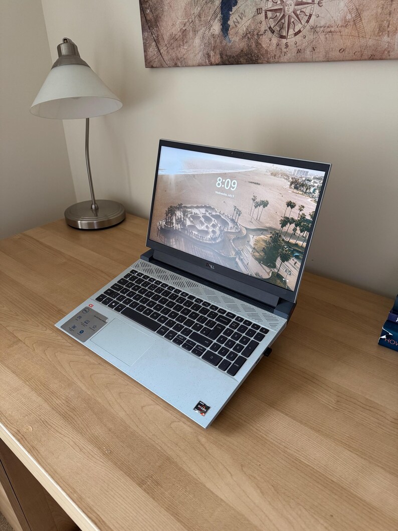 May include: A light blue laptop with a black keyboard is open on a wooden desk. The screen displays a beach scene with palm trees. A silver desk lamp and a decorative map are in the background.