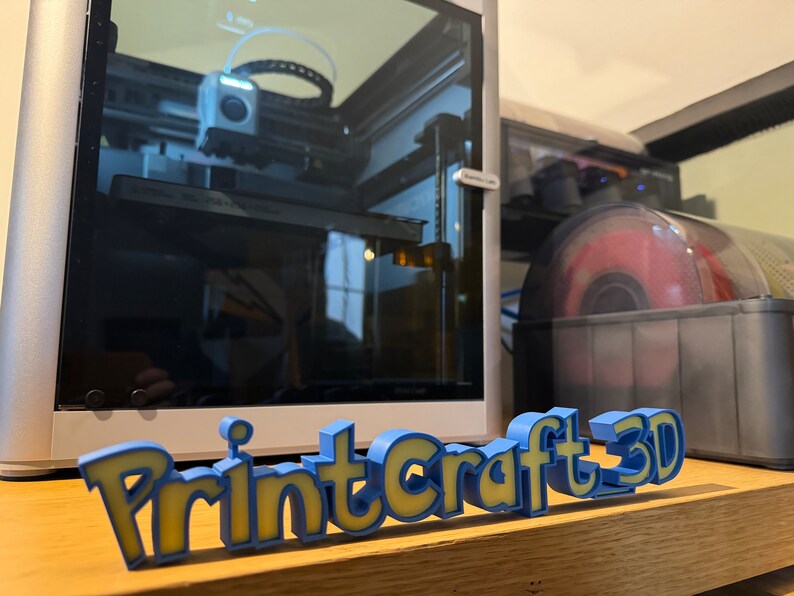 May include: A 3D printed sign that reads "PrintCraft 3D" in blue and yellow letters. The sign is sitting on a wooden surface in front of a 3D printer.