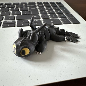 May include: A black 3D printed dragon figurine with yellow eyes. The dragon is lying on its belly with its wings spread out.
