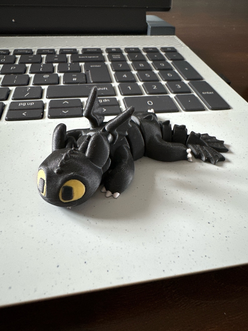 May include: A black 3D printed dragon figurine with yellow eyes. The dragon is lying on its belly with its wings spread out.