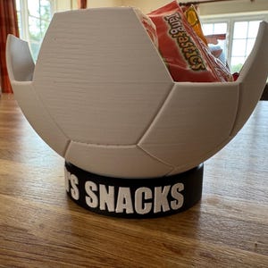 Op de afbeelding: Een witte, voetbalvormige snackkom met een zwarte basis. De basis leest "'S SNACKS" in witte letters. De kom is gevuld met oranje snackzakken.