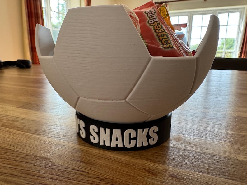 Op de afbeelding: Een witte, voetbalvormige snackkom met een zwarte basis. De basis leest "'S SNACKS" in witte letters. De kom is gevuld met oranje snackzakken.