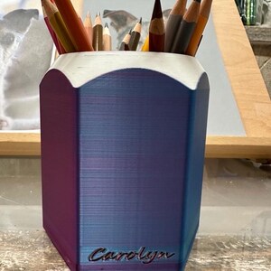 May include: A blue and purple pencil holder with a hexagonal shape and a white top. The holder has the name "Carolyn" engraved on the front.