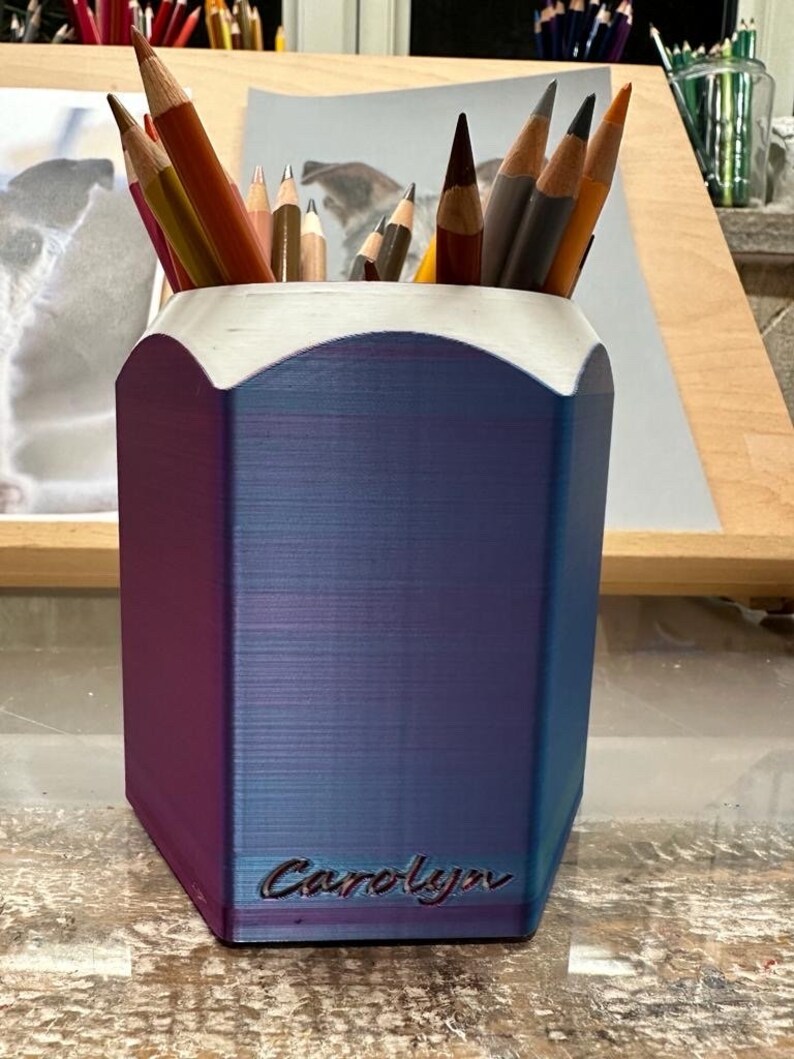 May include: A blue and purple pencil holder with a hexagonal shape and a white top. The holder has the name "Carolyn" engraved on the front.