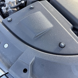 Può includere: Vista ravvicinata del vano motore di un'auto. L'immagine mostra una copertura del motore in plastica nera con tre elementi di fissaggio neri. I componenti del motore sono visibili sullo sfondo.