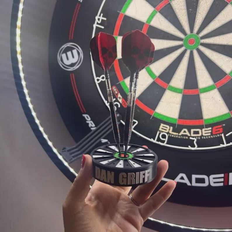 May include: A set of three darts with red skull-patterned flights, held in a black and green holder. The holder is inscribed with the name "DAN GRIFF". A dartboard is in the background.