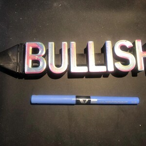 May include: A 3D printed word sign that reads "BULLISH" in iridescent pink, blue, and white letters. The sign is attached to a black, cylindrical base.