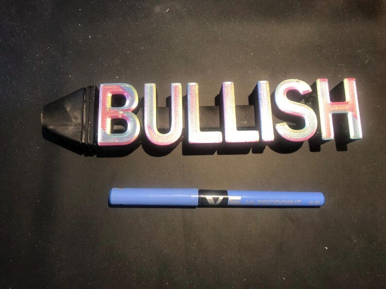 May include: A 3D printed word sign that reads "BULLISH" in iridescent pink, blue, and white letters. The sign is attached to a black, cylindrical base.