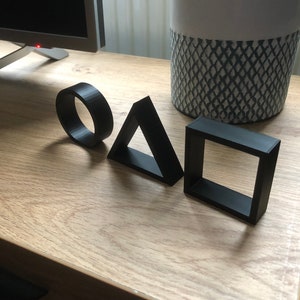 May include: Three black geometric shapes, a circle, a triangle, and a square, are arranged in a row on a wooden surface.