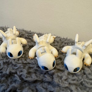 May include: Three white 3D printed figurines of a dragon with blue eyes. The dragons are lying on a gray fluffy surface.
