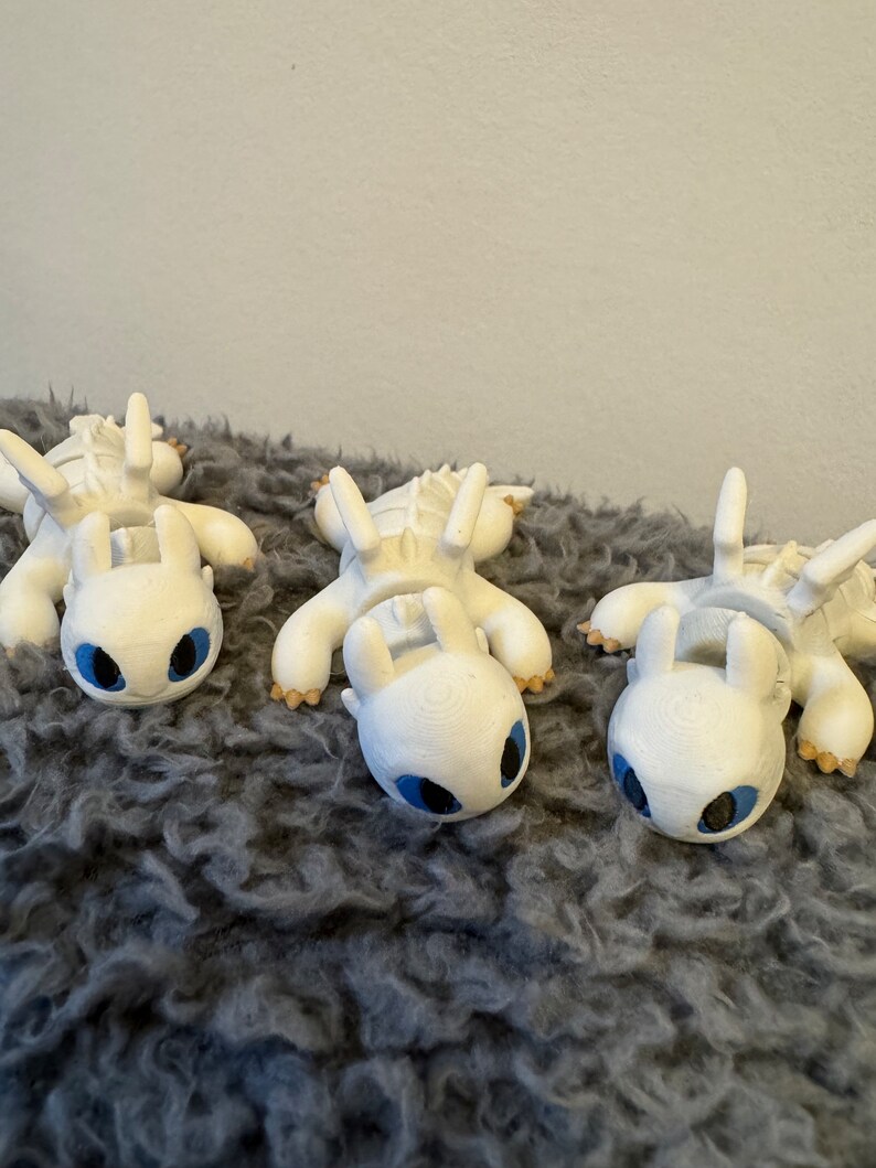 May include: Three white 3D printed figurines of a dragon with blue eyes. The dragons are lying on a gray fluffy surface.